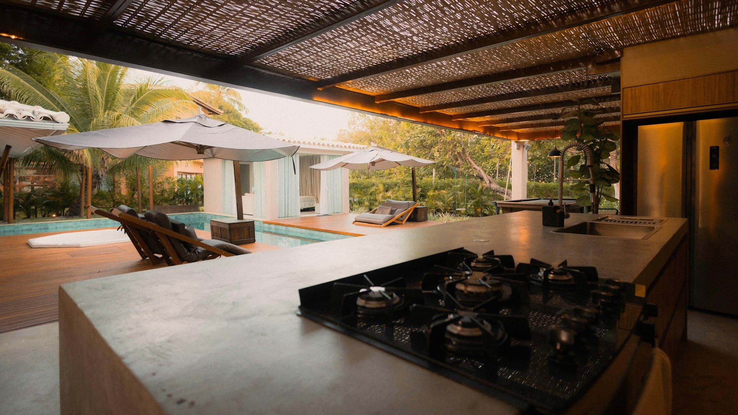 Outdoor kitchen with a gas cooktop and stone counters under a pergola, facing the pool