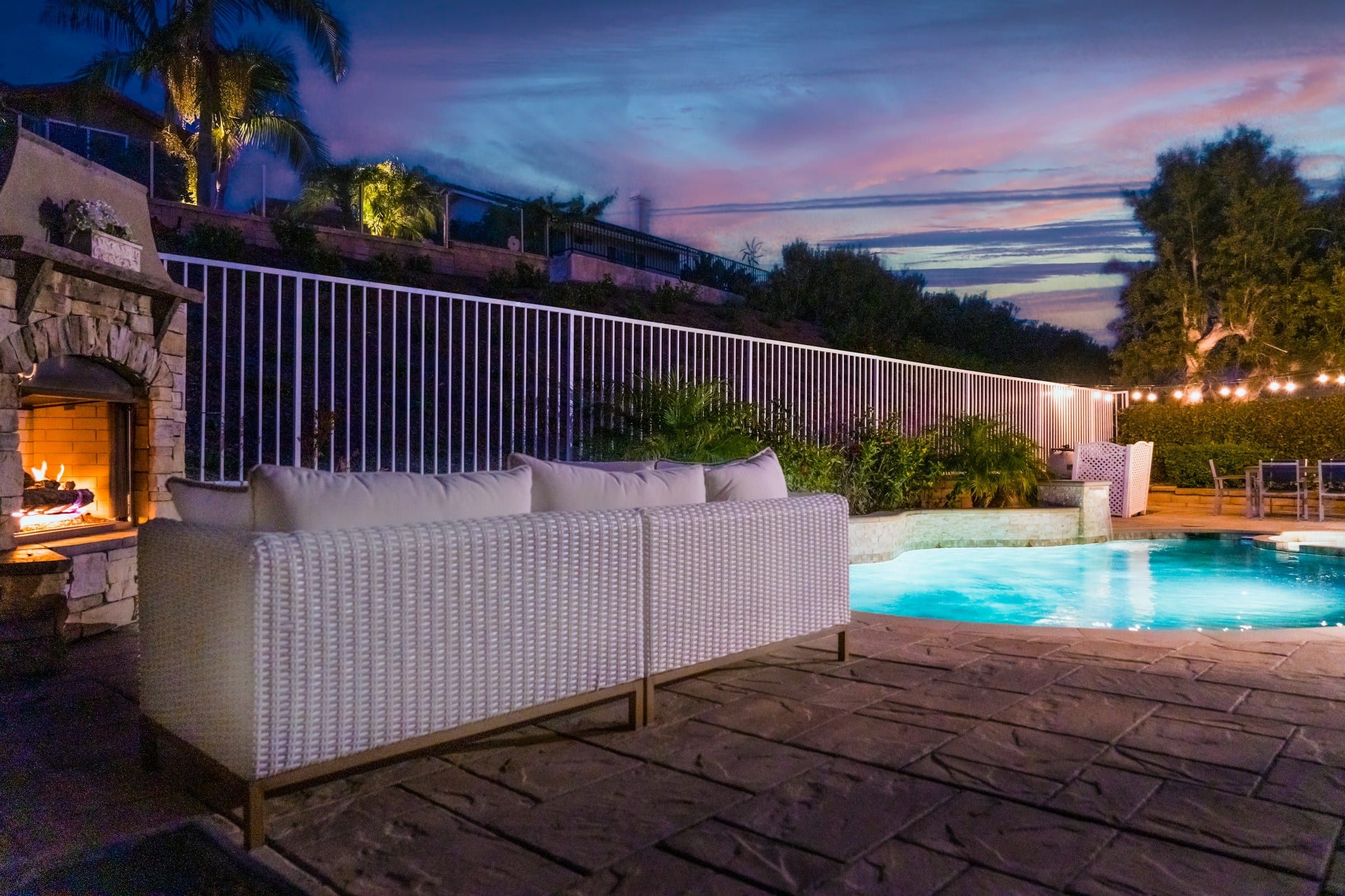 Backyard pool at dusk with a stone outdoor fireplace, lounge seating and string lights